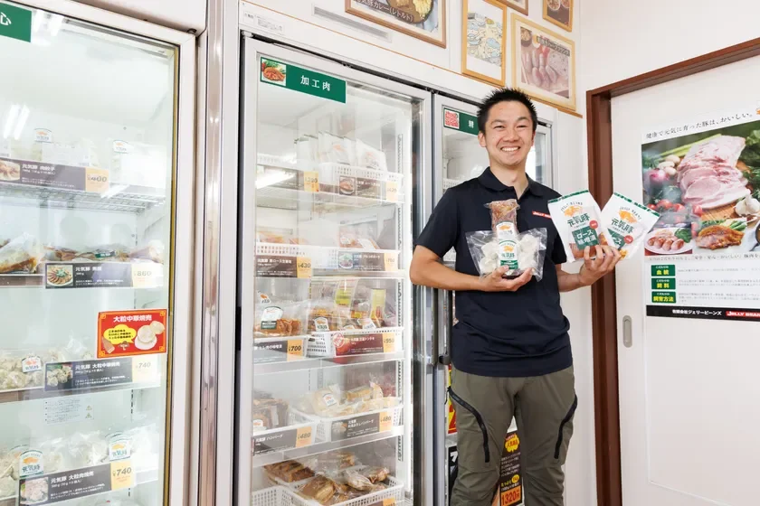 冷凍食品が並ぶショーケースの前で、笑顔の男性が豚肉の加工品を手に持っています