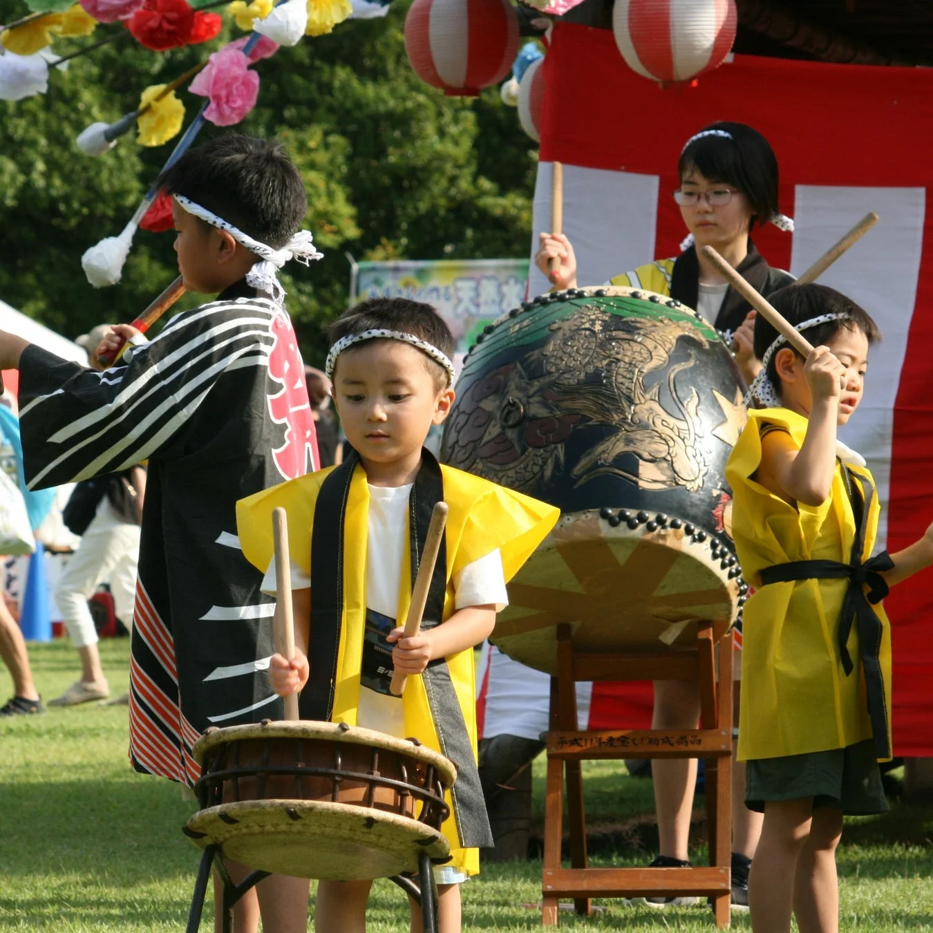 日本の祭りで、数人の子供たちが伝統的な衣装を身につけ、太鼓を演奏している様子です。色とりどりの提灯や飾りが背景に見え、賑やかな雰囲気が伝わります。
