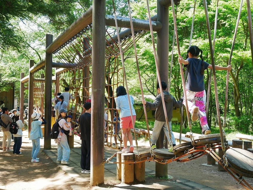 緑豊かな公園のアスレチックで、子供たちがロープや木製の遊具を使って楽しそうに遊んでいます。大人たちも周囲で見守り、家族連れで賑わう休日の様子がうかがえます。
