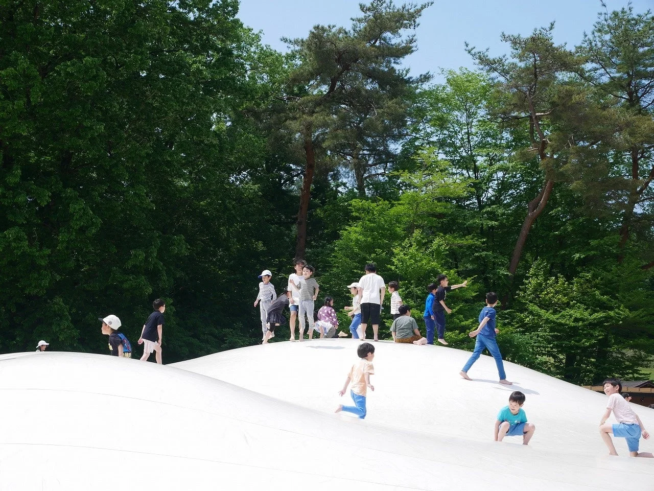 森に囲まれた広々とした白いふわふわドームで、たくさんの子供たちが元気に飛び跳ねたり走ったりして遊んでいます。晴れた日の屋外での楽しいひとときを捉えた写真です。