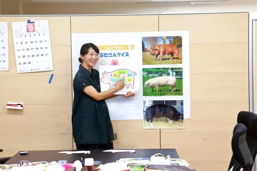 オフィスで女性が「ぶたさんクイズ」と題されたポスターを指し示し、笑顔で説明