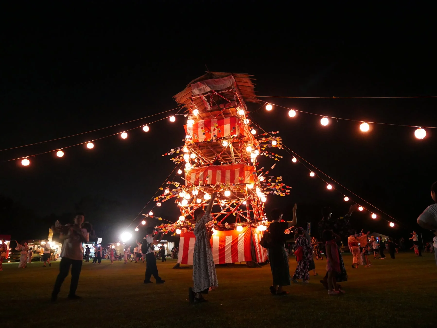 夜の広場で開催されている日本の伝統的な夏祭りの様子です。中央には提灯や紅白幕で飾られたやぐらが設置され、その周りには多くの人々が集まり、盆踊りを楽しんでいます。会場には暖かな光を放つ裸電球が吊るされ、幻想的な雰囲気を醸し出しています。