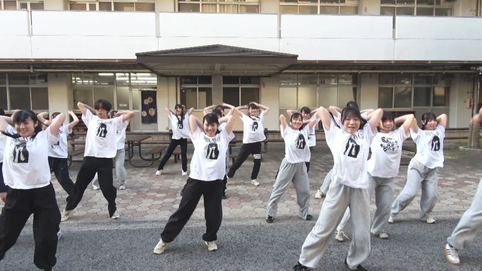 白いTシャツを着た多くの学生が屋外の校庭で一斉にダンスを練習しています。
